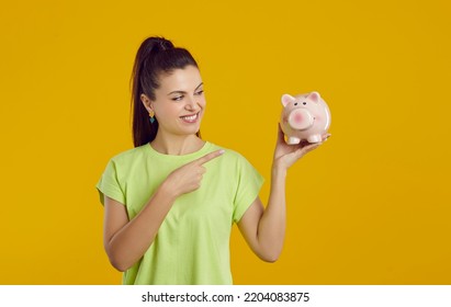 Smiling Woman Points To Piggy Bank In Her Hand, Hinting At Favorable Offer From Bank. Happy Caucasian Casual Woman Holding Piggy Bank In Form Of Pink Pig. Isolated On Vivid Yellow Background. Banner.