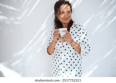 Smiling woman in a polka dot shirt holds a coffee cup, standing against a bright wall, enjoying a peaceful and sunny moment. - Powered by Shutterstock