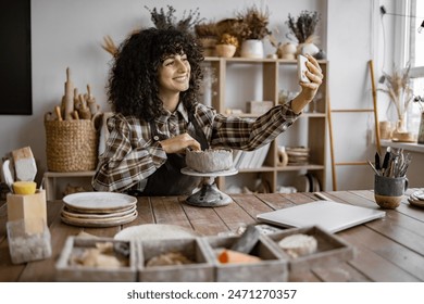 Smiling woman potter taking selfie while sculpting pottery on rotating wheel in cozy art studio - Powered by Shutterstock