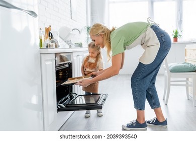 smiling woman putting baking sheet in electric oven near little daughter