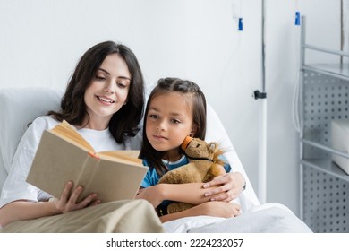 Smiling woman reading book near daughter with toy on bed in hospital bed - Powered by Shutterstock