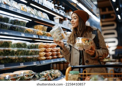 Smiling woman reading label on food package while buying groceries from refrigerated section in supermarket.