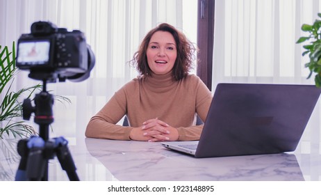 Smiling woman recording her video blog. Woman blogger videotapes her vlog at home.  Adult woman speaks in front of a video camera for her blog channel. Vlogger makes online streaming using smartphone