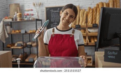 Smiling woman in a red apron holds tongs at a bakery with bread shelves and pastries behind the counter - Powered by Shutterstock