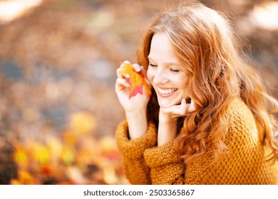 A smiling woman with red hair playfully holds an autumn leaf near her face, wearing a cozy sweater, enjoying the outdoor autumn atmosphere with colorful, blurred foliage in the background
 - Powered by Shutterstock