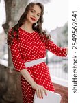 Smiling woman with red lipstick and curly hair wearing red polka dot dress and holding white clutch bag poses in urban setting