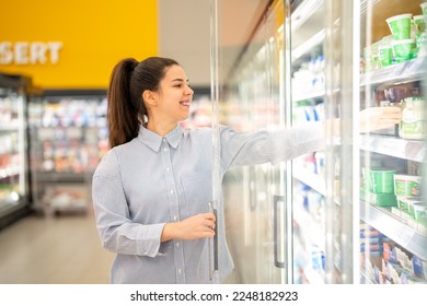 Smiling woman at the refrigerated section in a supermarket, opening the freezer and reaching for a diary product. - Shutterstock ID 2248182923