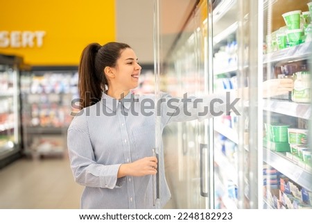 Smiling woman at the refrigerated section in a supermarket, opening the freezer and reaching for a diary product.