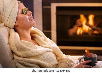 Smiling Woman Relaxing At Home With Kiwifruit Facial Mask And Tea Mug, Sitting In Bathrobe In Front Of Fireplace.