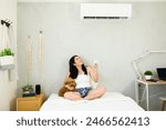 Smiling woman with a remote control enjoys the cooling breeze from a mini split air conditioning unit on a hot summer day