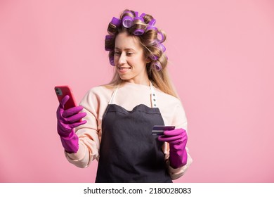 Smiling Woman With Rollers On Head Poses Against Pink Background Holds Credit Card In Hand Protected By Rubber Glove, Orders Cleaning Accessories, Pays In App On Phone.