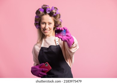 Smiling Woman With Rollers On Head Poses Against Pink Background Holds Credit Card In Hand Protected By Rubber Glove, Orders Cleaning Accessories, Pays In App On Phone.