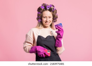 Smiling Woman With Rollers On Head Poses Against Pink Background Holds Credit Card In Hand Protected By Rubber Glove, Orders Cleaning Accessories, Pays In App On Phone.