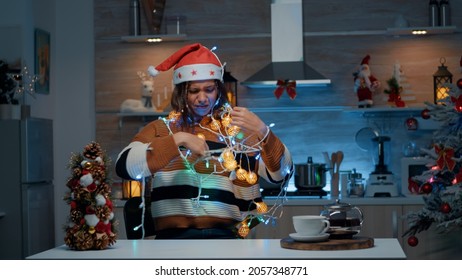 Smiling woman with santa hat tangled in christmas lights string while doing decor preparations in kitchen. Young person having fun indoors with festive decorations and ornaments - Powered by Shutterstock