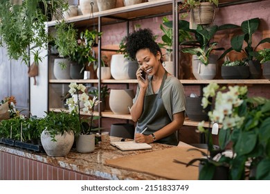 Smiling woman seller in botanical shop talking on smartphone. African american botanist writing client order in notebook during conversation on mobile phone in store. Young black small business owner. - Shutterstock ID 2151833749