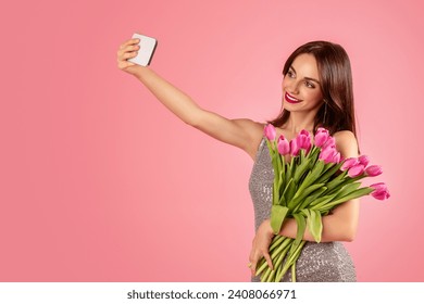 Smiling woman in a sequined dress taking a selfie with her smartphone, clutching a bunch of fresh pink tulips, against a soft pink background. Spring holiday celebration, photo app