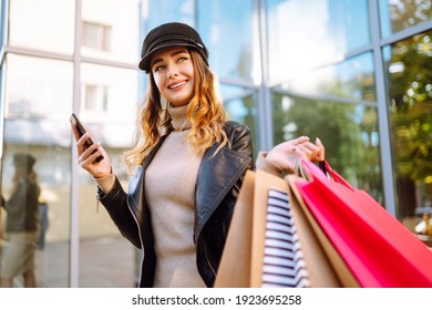 Smiling Woman With Shopping Bags Using Her Phone. Young Woman After Shopping On The City Street. Purchases, Black Friday, Discounts, Sale Concept. Online Shopping Concept.