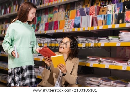 smiling woman showing new colorful copybooks to daughter in stationery store