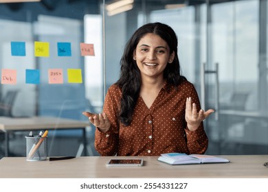 A smiling woman sits in a modern office, engaging in a discussion. She displays open body language, conveying confidence and positivity.