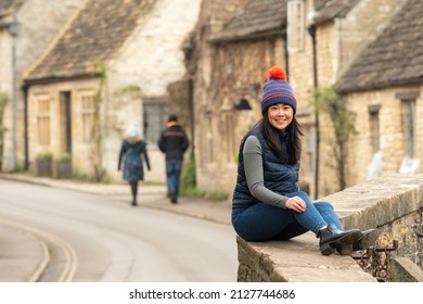 Smiling woman sits on a bridge in front of a charming street in the traditional English village of Castle Combe, Wiltshire, with Cotswold stone houses in the background. Tourist on UK winter holiday. - Powered by Shutterstock