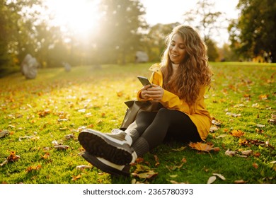 A smiling woman sits outdoors on the lawn among yellow autumn leaves with a phone in her hands. A female uses a smartphone in an autumn park, enjoying nature. - Powered by Shutterstock