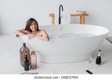 Smiling woman sitting in bathtub near champagne and smartphone on floor - Powered by Shutterstock