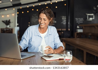 Smiling woman sitting in cafeteria holding coffee mug and working on laptop. Businesswoman checking email on laptop. Beautiful middle aged woman and using laptop at cafe while drinking a cup of tea.