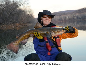 A smiling woman sitting in a canoe in a blue and gold dry suit holding a large brown flathead catfish horizontally  on a mirrored river in winter