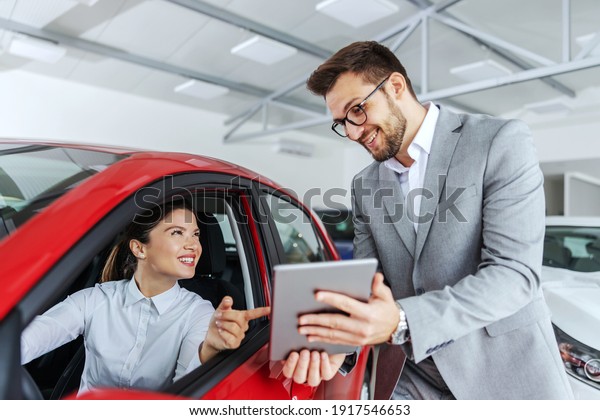 Smiling woman sitting in car and pointing at\
tablet car seller holding. She is picked right car for her she saw\
online. Car salon\
interior.