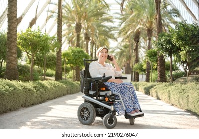 a smiling woman sitting in an electric wheelchair with disability enjoys a sunny day in the garden of the city park talking on her mobile phone - Powered by Shutterstock