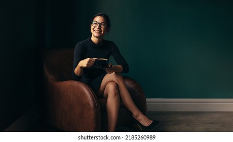 Smiling woman sitting on chair and holding coffee cup. Happy asian female enjoying coffee while sitting on armchair. - Shutterstock ID 2185260989