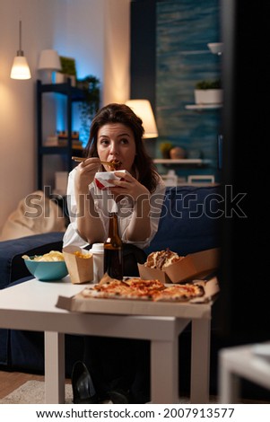 Smiling woman sitting on couch eating tasty chinese food during fast-food home delivered in living room. Caucasian female watching at television enjoying chopstick. Takeaway food delivery