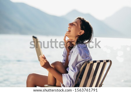 Smiling woman sitting on deck chair by the sea using tablet on a sunny day