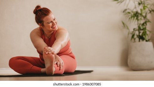 Smiling woman sitting on fitness mat and stretching leg. Fit female doing warmup workout at fitness studio. - Shutterstock ID 1986307085
