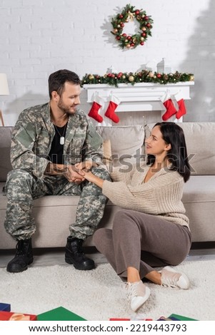 smiling woman sitting on floor and holding hands of husband in camouflage in living room with christmas decor