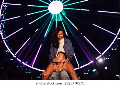 Smiling woman sitting on the shoulders of her boyfriend and looking at him. Young couple at night against ferris wheel with colorful lights.