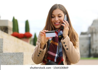 Smiling Woman Sitting Outdoors Looking At Number On Credit Card And Confirm Purchase Via Telephone Call To Customer Service. Positive Girl Making Payment Via Smartphone Conversation Outside.