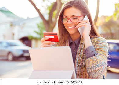 Smiling Woman Sitting Outdoors Talking On Mobile Phone Making Online Payment On Her Tablet Computer Outside On A Sunny Autumn Day 