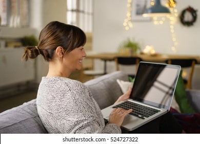 Smiling Woman Sitting Surfing The Internet On A Laptop Computer On A Sofa At Home, Side Rear View With The Screen With Reflections Visible