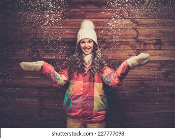 Smiling Woman In Ski Jacket And White Hat Standing Against Wooden House Wall Under Snow 