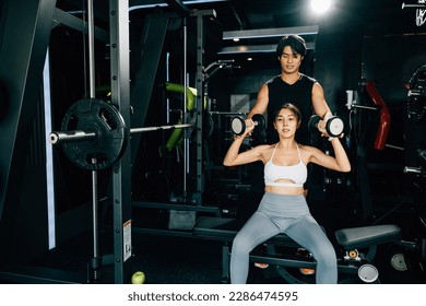 Smiling woman in sportswear exercising on a bench with dumbbells, guided by her personal trainer for a safe and effective workout, technique of exercise in gym dark gym background - Powered by Shutterstock