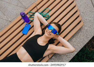 Smiling woman in sporty attire relaxing on a bench with a water bottle, jump rope, and smartphone, highlighting fitness and relaxation - Powered by Shutterstock