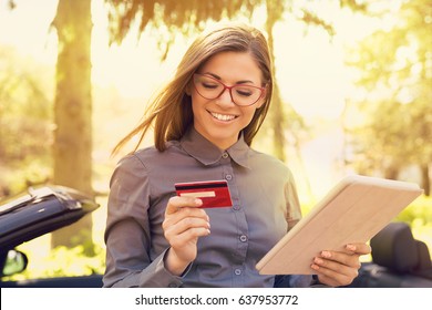 Smiling Woman Standing By Her New Car Making Online Payment On Her Tablet Computer Outside On A Sunny Summer Day  