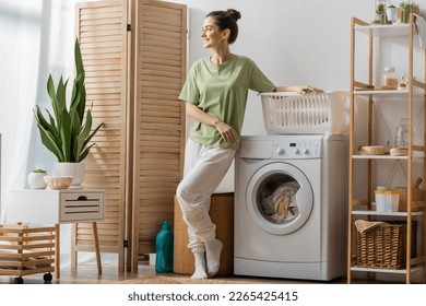 Smiling woman standing near basket and washing machine at home - Powered by Shutterstock