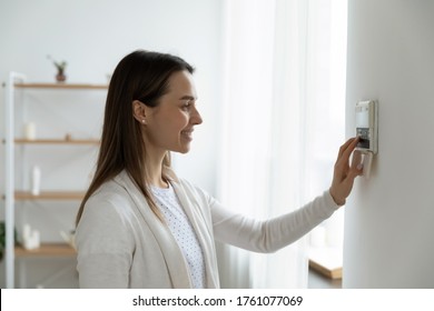Smiling woman standing near wall-mounted device adjusting degrees in living room set comfortable temperature using thermostat home heating system. Owner of modern smart house, energy saving concept - Shutterstock ID 1761077069