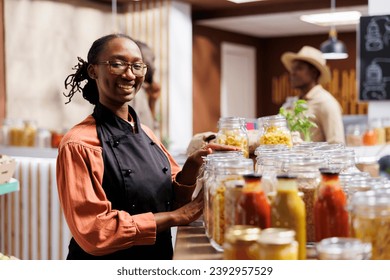 Smiling woman standing next to a wooden shelf filled with bulk food glass containers. These sustainable products are neatly organized for customers who prefer a zero-waste shopping experience. - Powered by Shutterstock