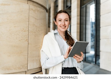 A smiling woman stands outside holding a tablet, dressed in business casual attire with an urban building backdrop. - Powered by Shutterstock