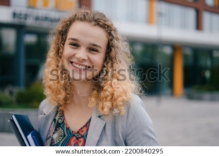 Smiling woman student holding books and a laptop, looking at camera standing outdoors university. Happy confident young business woman, freelance employee
