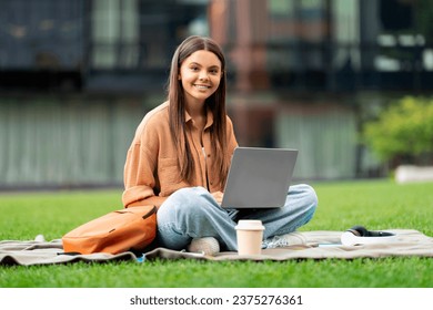 A smiling woman student uses her laptop at a university campus. Seated on green lawn, she types and occasionally looks around, absorbing the lively campus atmosphere while focused on her screen - Powered by Shutterstock