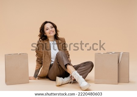 smiling woman in suede jacket and brown pants sitting near shopping bags on beige background
